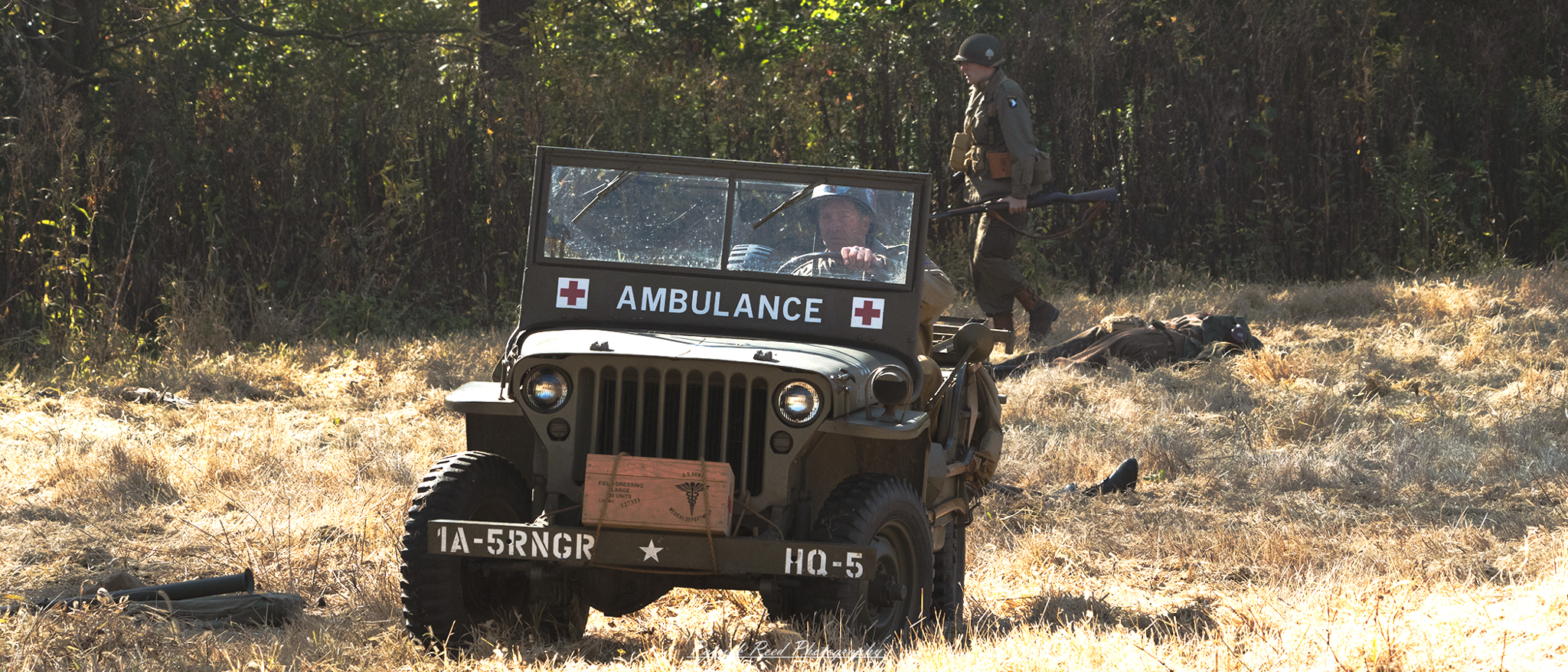 A World War II ambulance jeep parked on a battlefield, featuring a distinctive red cross emblem on its side. The sturdy vehicle is equipped with stretchers and medical supplies, designed to transport wounded soldiers to safety. Dust and dirt cover the rugged tires, reflecting the harsh conditions of the war zone. The jeep's utilitarian design and compact frame highlight its role as a vital medical lifeline during the conflict, with the open battlefield and scattered debris visible in the background.